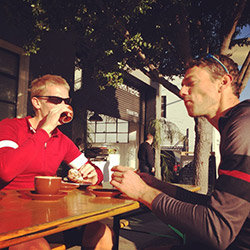 Two men in bicycle jerseys sitting outside at table having coffee
